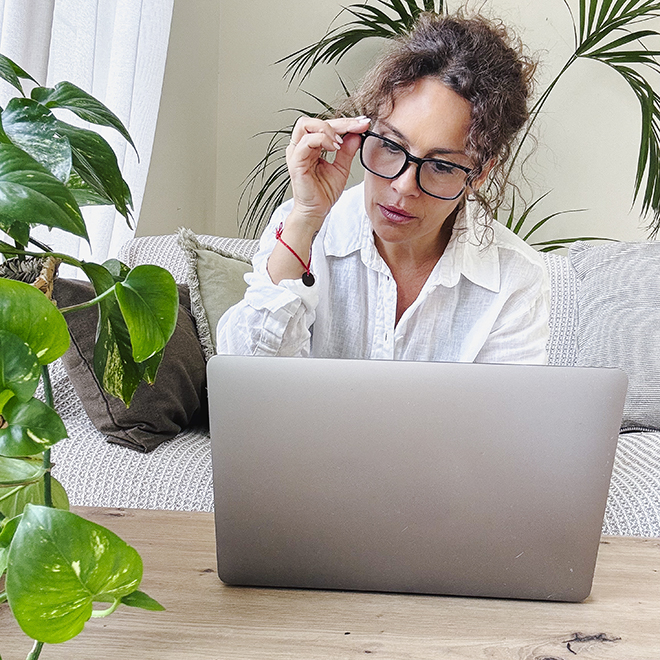 Woman adjusting her spectacles while looking intently at a laptop screen, working remotely from a comfortable home office setup with green plants creating a peaceful atmosphere